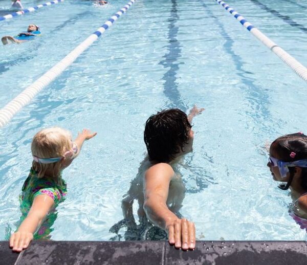 An adult teaches two children how to swim in a pool, with the children holding onto the pool edge and other people swimming in designated lanes.