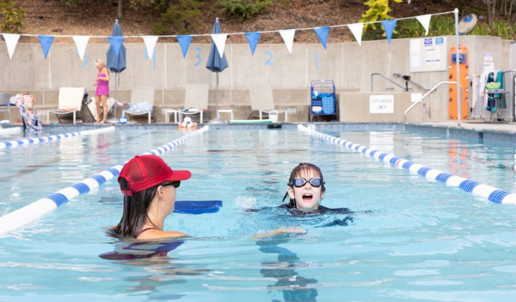 A swim instructor wearing a red cap assists a child with goggles in an outdoor pool lane; other people are visible in the background.