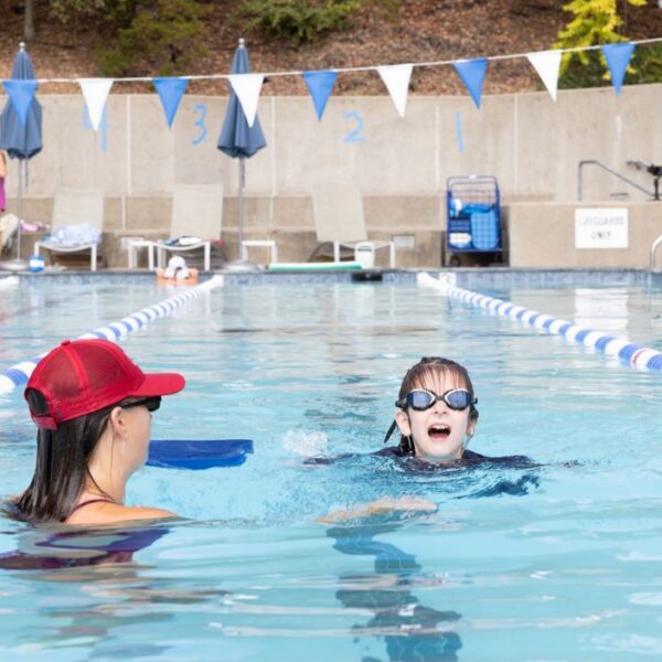 A swim instructor wearing a red cap assists a child with goggles in an outdoor pool lane; other people are visible in the background.
