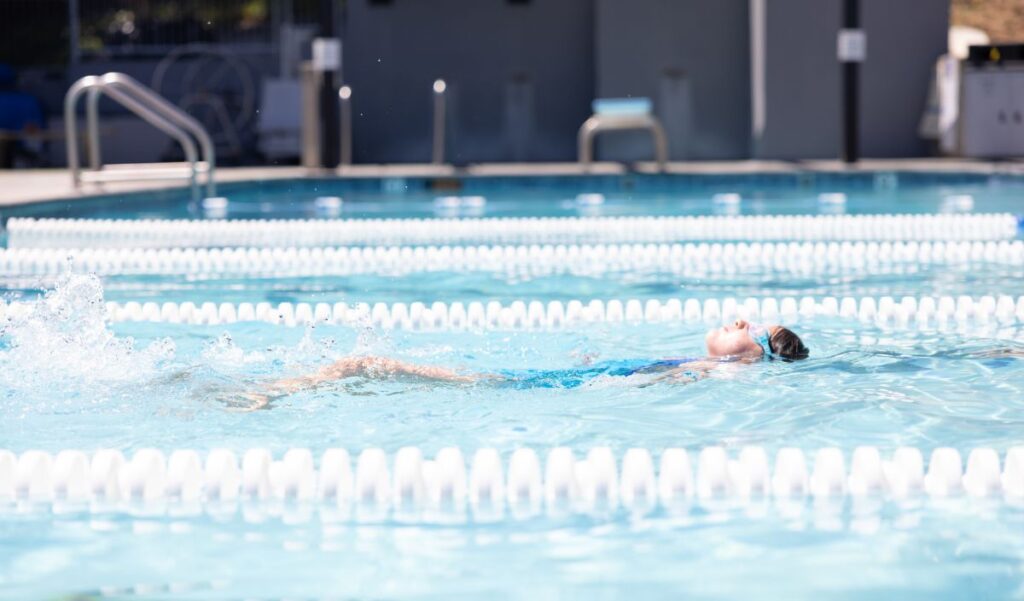 A person wearing goggles and a swim cap swims backstroke in a clear outdoor pool, creating splashes as they move through a swim lane.