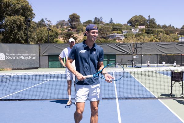 Two men stand on an outdoor tennis court; the man in front holds a racket and smiles, while the man behind him stands near the net.