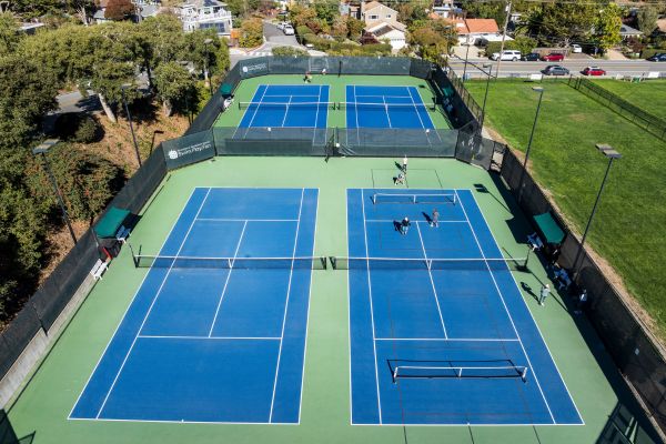 Aerial view of four outdoor blue tennis courts with people playing; residential area and green lawn visible in the background.
