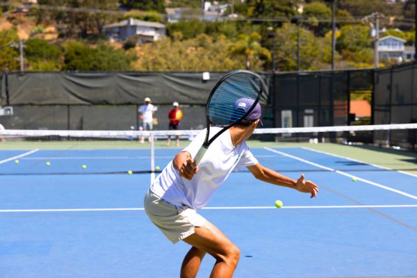 A tennis player in a white shirt and shorts prepares to hit a backhand shot on an outdoor court, with several tennis balls scattered around.