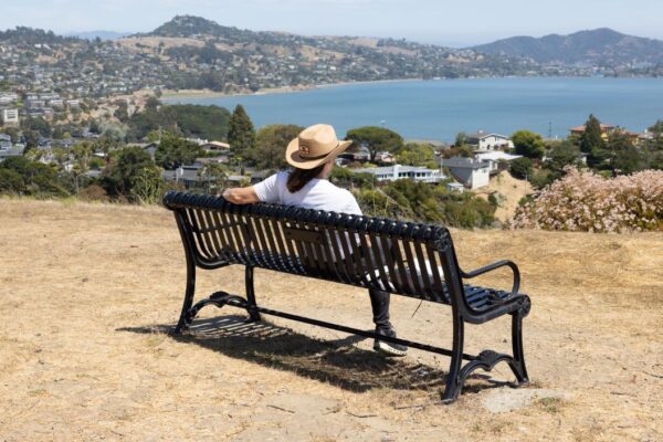 A person wearing a hat sits on a bench overlooking a bay, with houses, hills, and water visible in the background under clear skies.