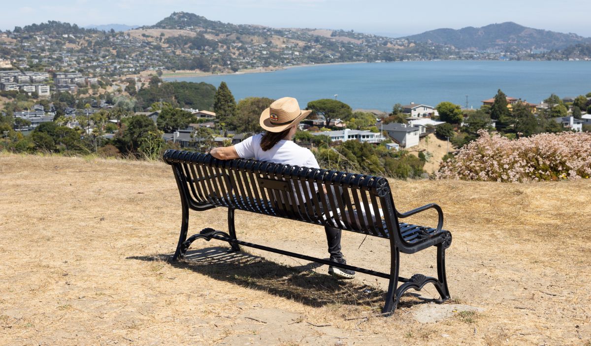 A person wearing a hat sits on a bench overlooking a bay, with houses, hills, and water visible in the background under clear skies.