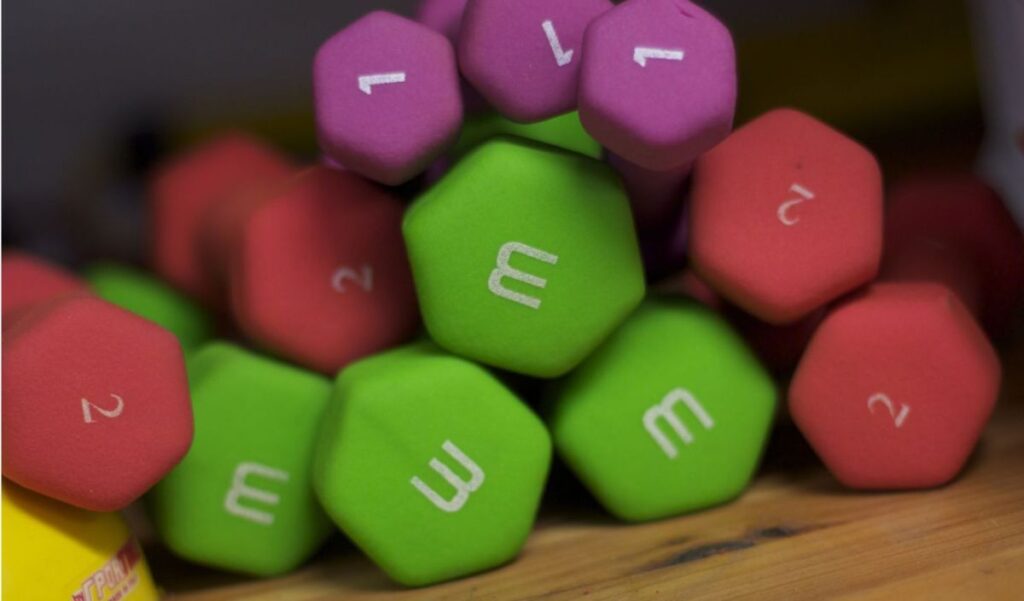 A pile of hexagonal dumbbells in green, red, and purple, labeled with white numbers and the letter "M," resting on a wooden surface.