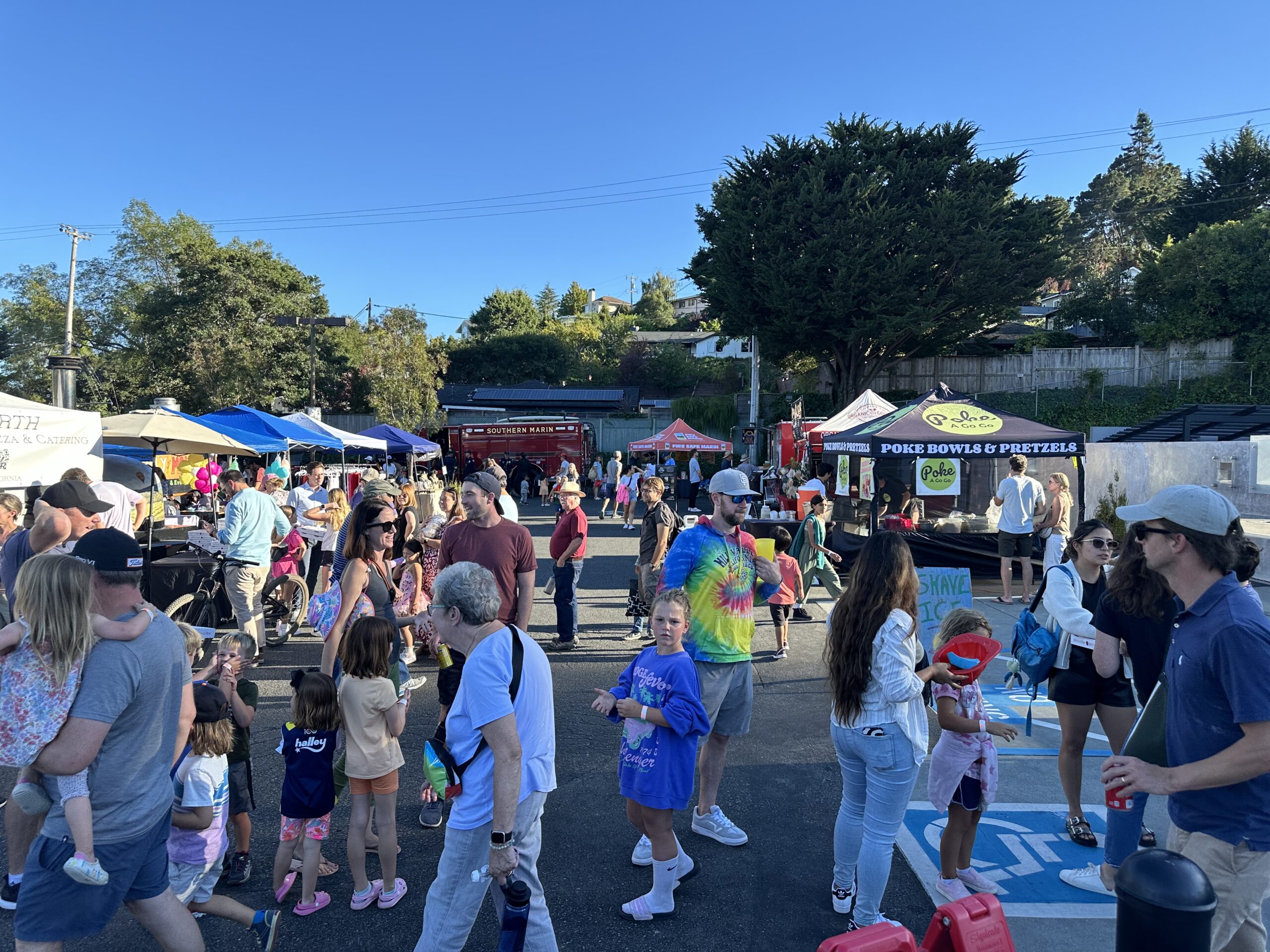 A crowded outdoor market with people walking, shopping at vendor stalls, and children playing on a sunny day.