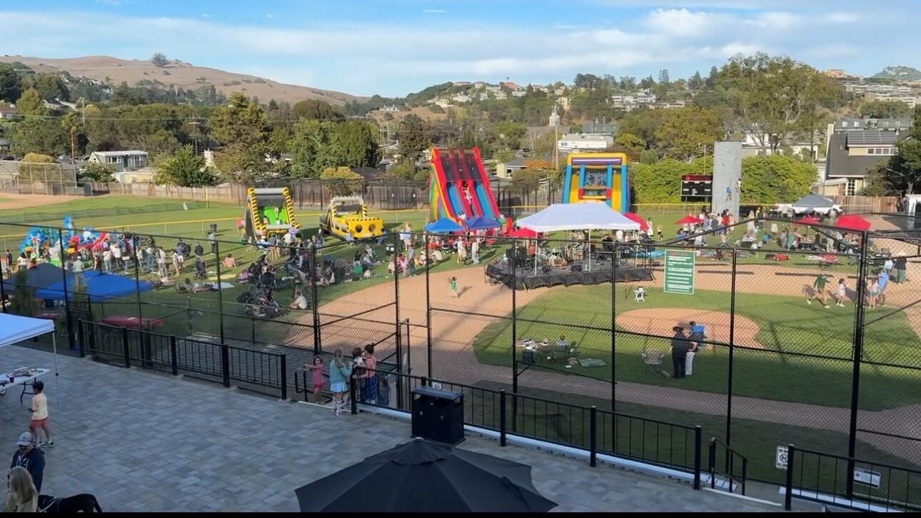 A community event at a baseball field features people, bounce houses, tents, and activity stations on a sunny day, with hills and houses in the background.