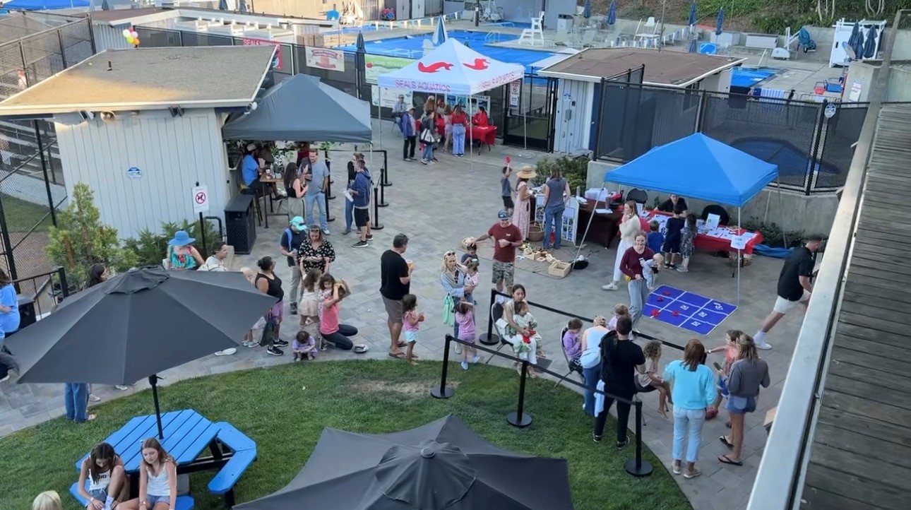 A group of people, including children and adults, gather outdoors under tents and umbrellas, participating in games and activities at a community event.