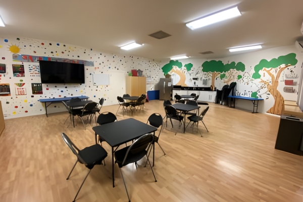 A classroom with tables and chairs, wall art featuring trees and educational designs, light wood flooring, and bright overhead lighting.