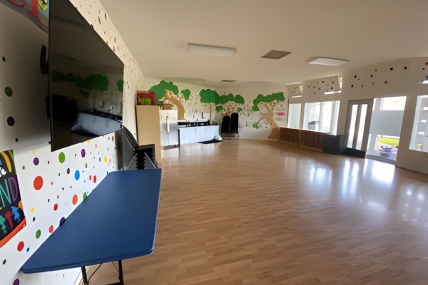A brightly lit, empty classroom with wood flooring, colorful wall art, tables, shelves, and large windows letting in natural light.