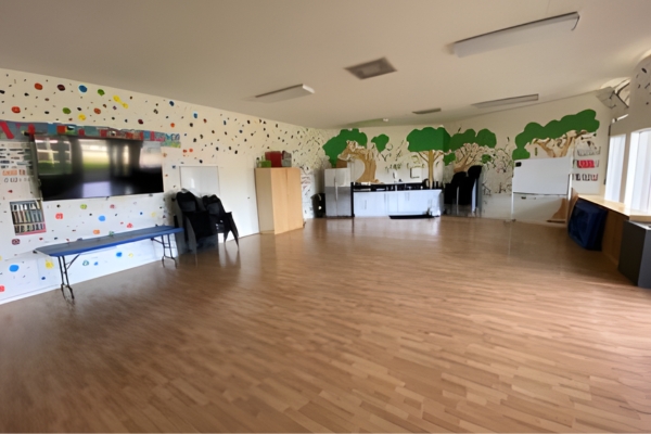 A spacious room with wood flooring, wall murals of trees, chairs, cabinets, a bench, a TV, and large windows letting in natural light.