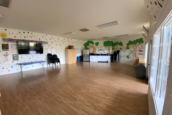 A spacious room with wooden floors, wall murals of trees, a table, stacked chairs, cabinets, a TV, and windows letting in natural light.