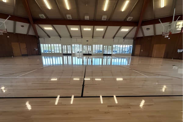 Indoor basketball court with polished wooden floor, high ceiling, visible basketball hoops, and large windows letting in natural light.