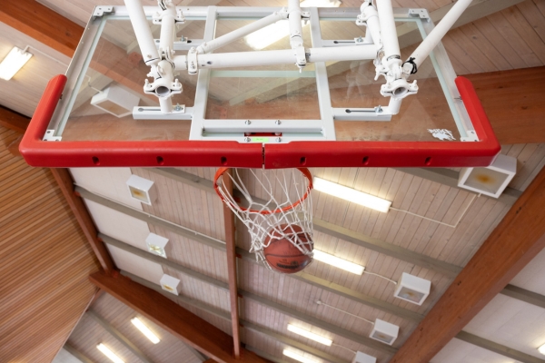 A basketball going through a net attached to a glass backboard in an indoor gym with ceiling lights and beams visible.