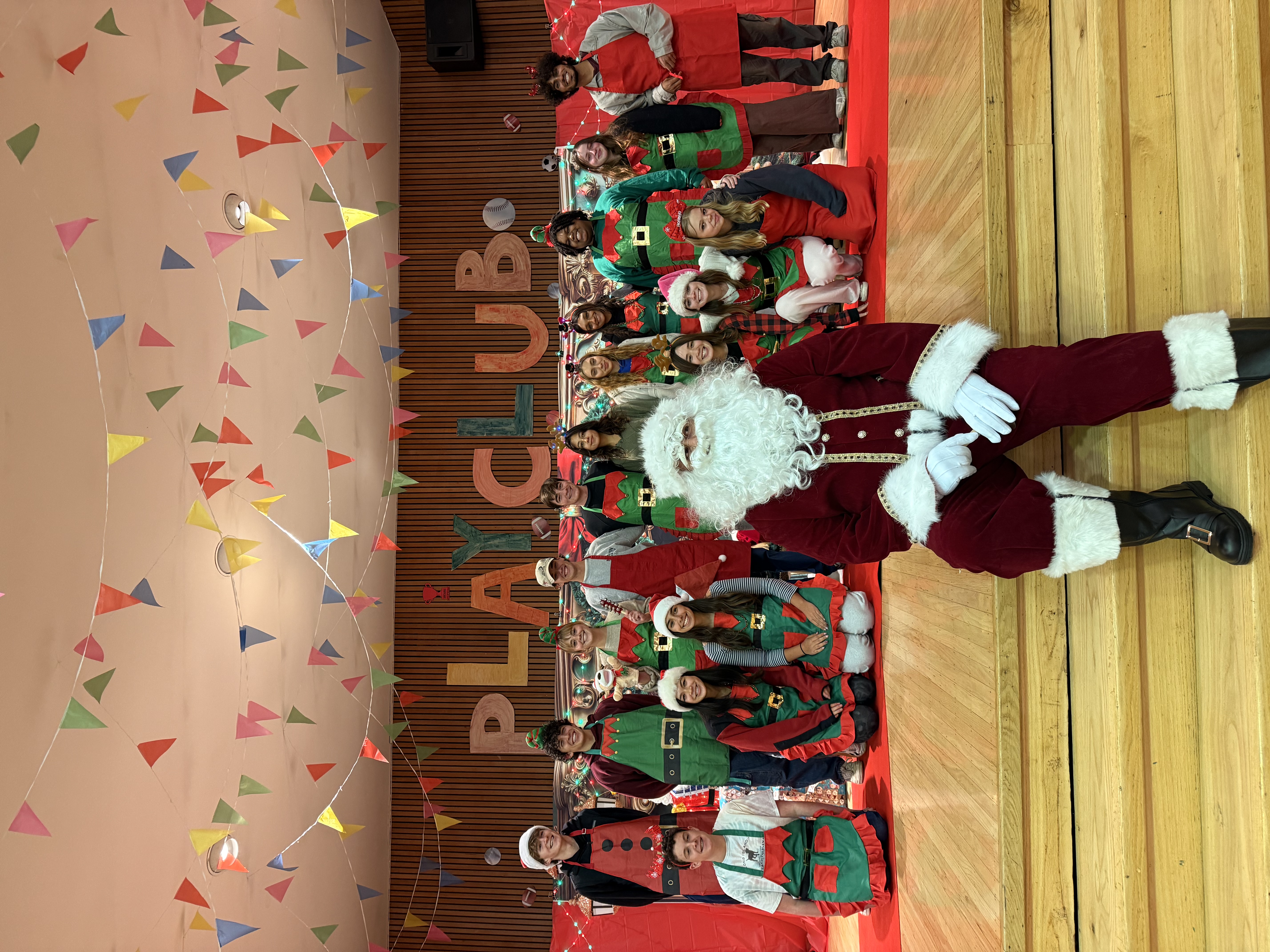 A person dressed as Santa Claus sits on a stage in front of a group of people in holiday costumes, with colorful banners and "PLAY CLUB" sign in the background.