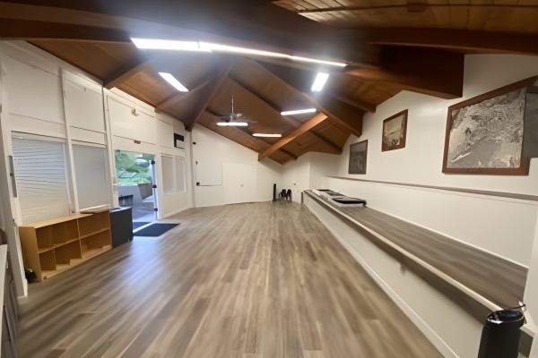 A spacious, empty room with wooden floors, high wooden ceilings, framed photos on the wall, shelves, and fluorescent lighting.