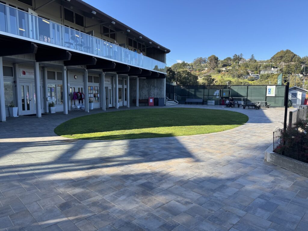 A modern school courtyard with a circular grassy area, paver stone ground, two-story building, and outdoor seating under a clear sky.