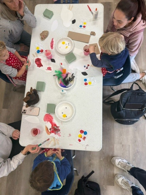 Children and adults sit around a table painting and doing crafts with various art supplies, including paint, feathers, and paper.