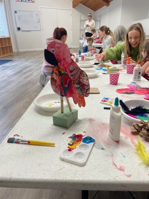Children and adults sit around a long table working on colorful craft projects, including a painted bird sculpture, with paint, brushes, and glue visible on the table.