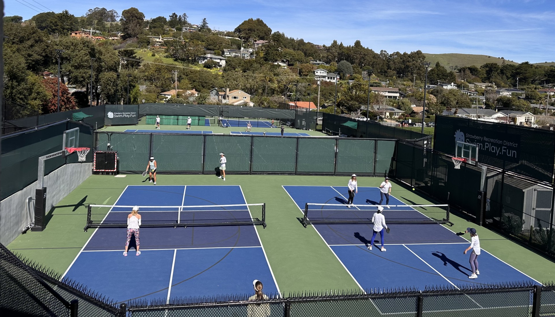 People play pickleball and tennis on outdoor courts surrounded by fencing, with houses and hills visible in the background under a clear sky.