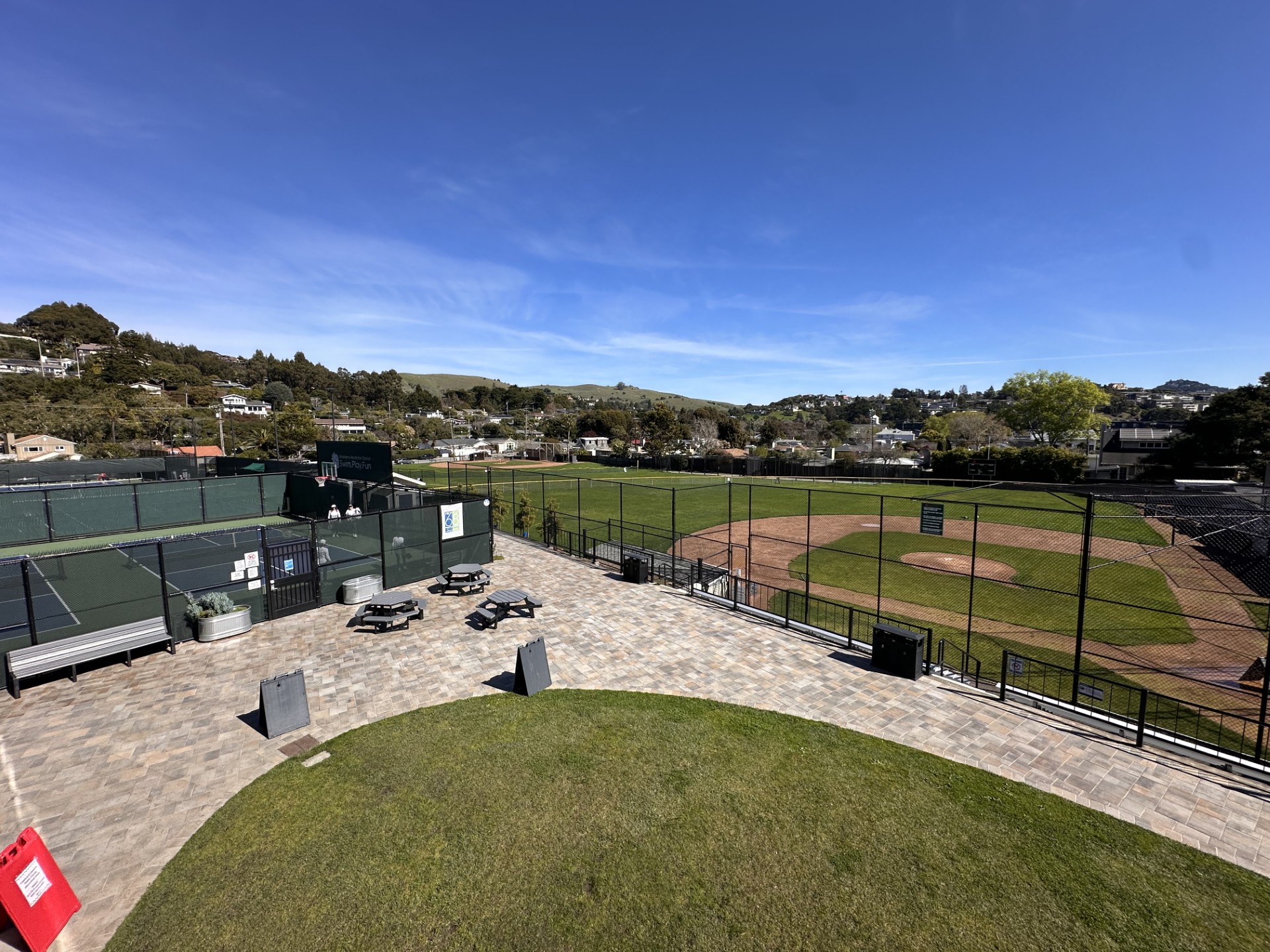 View of a baseball field with green stands, empty bleachers, picnic tables, and a paved area in the foreground under a clear blue sky.
