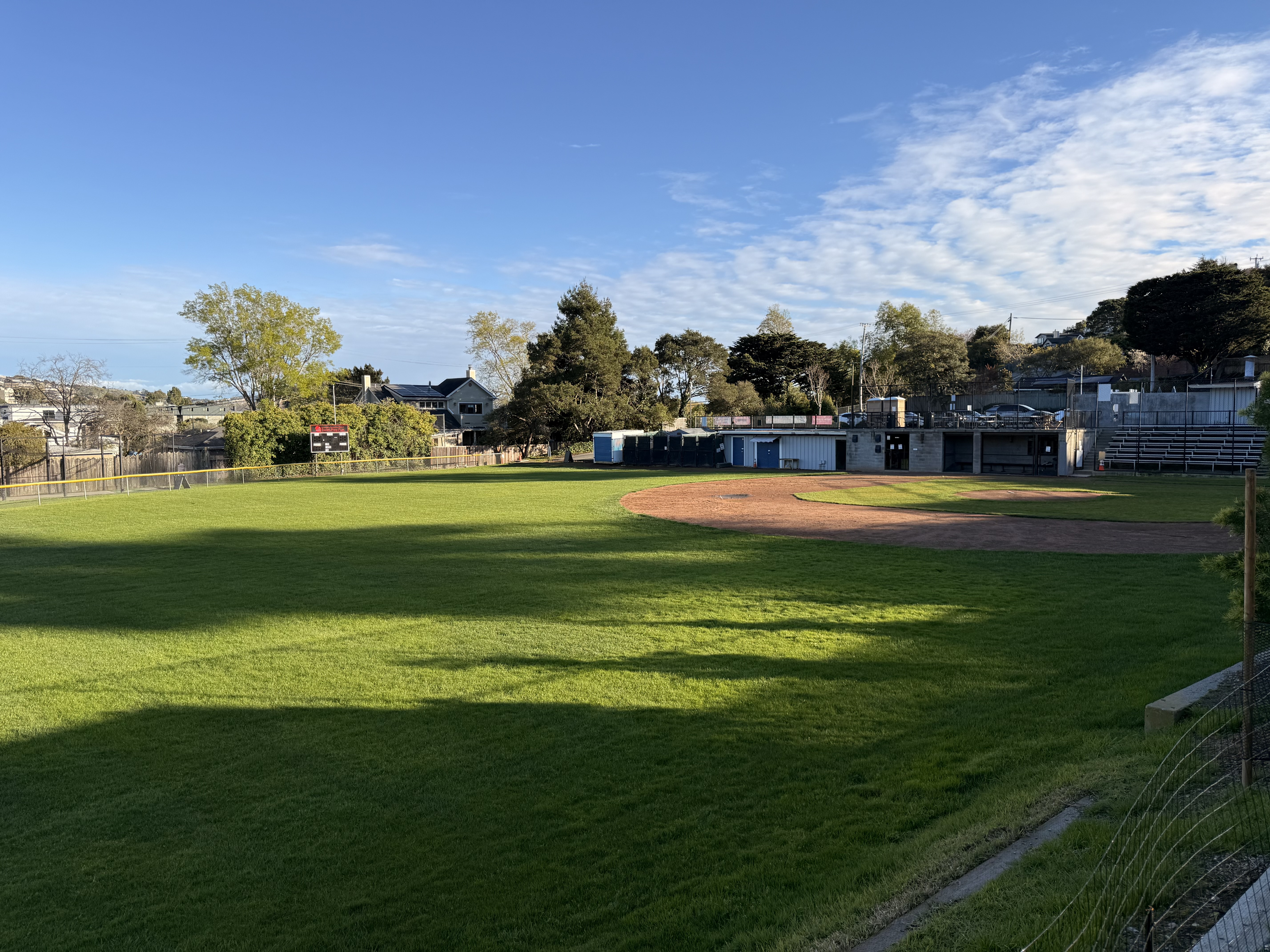 A baseball field with green grass, dirt infield, empty bleachers, and buildings in the background under a partly cloudy sky.