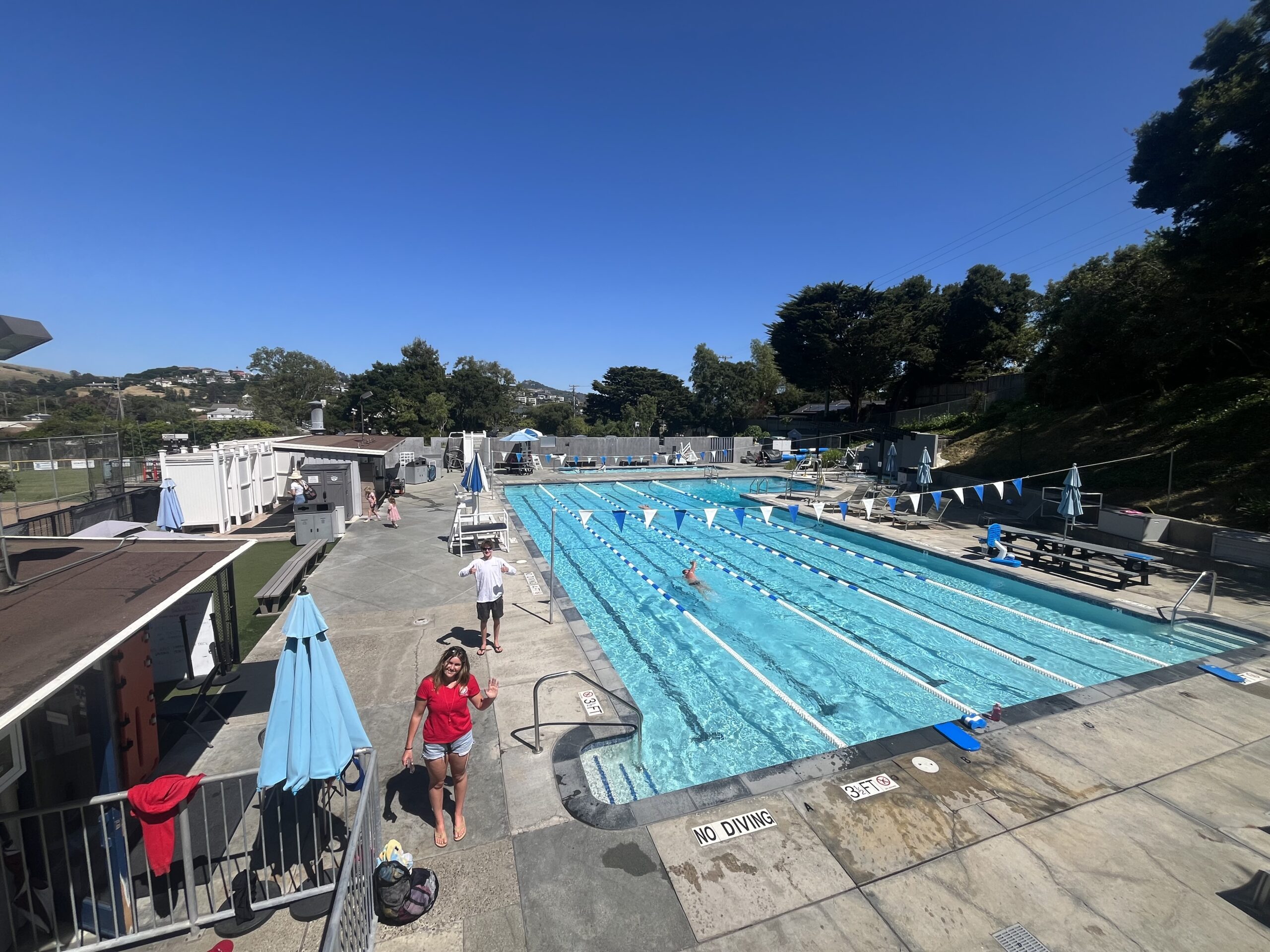 Outdoor swimming pool with marked lanes; a few people are swimming while others stand or walk around the pool deck on a sunny day.