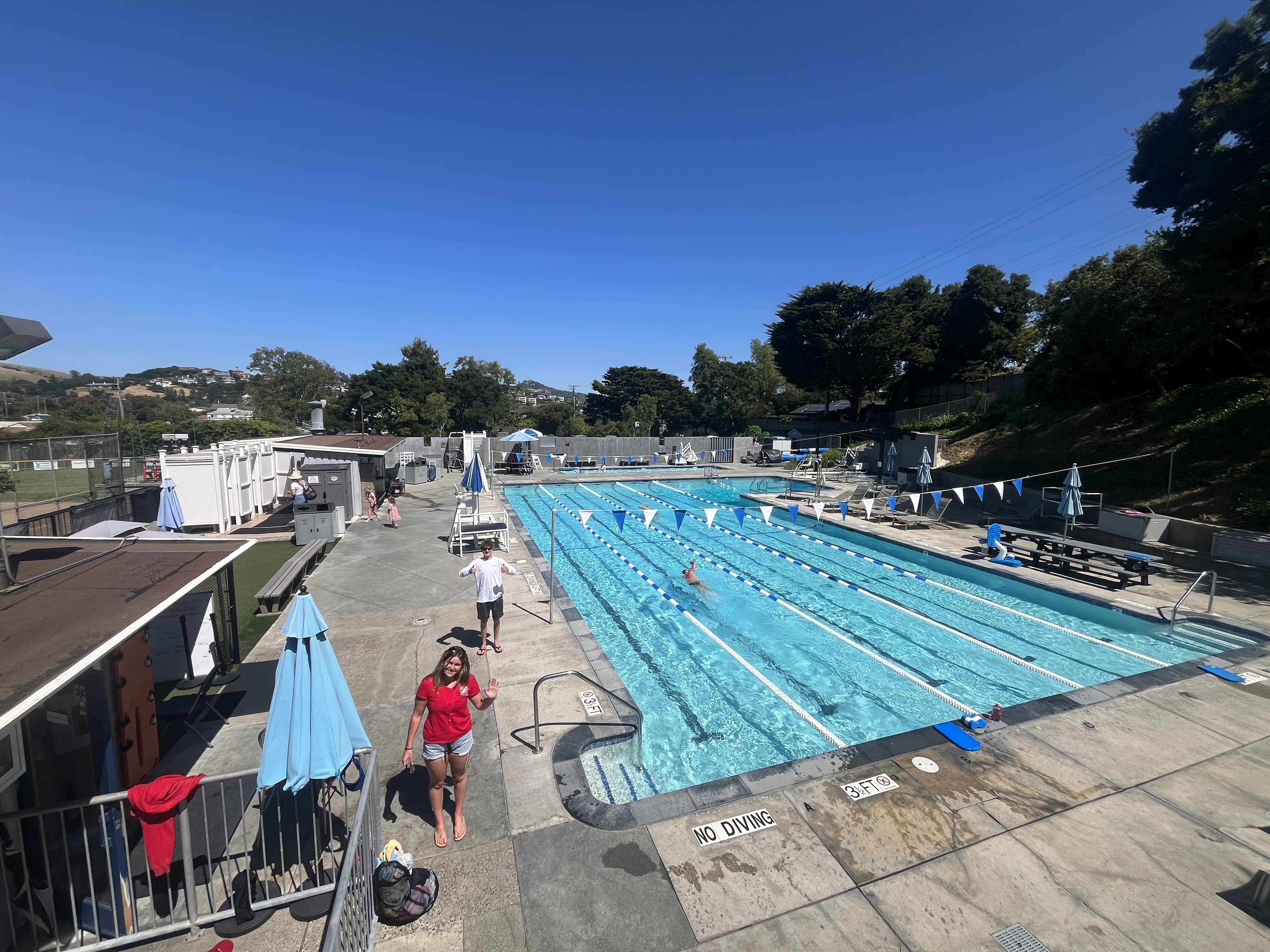 Outdoor swimming pool with marked lanes; a few people are swimming while others stand or walk around the pool deck on a sunny day.