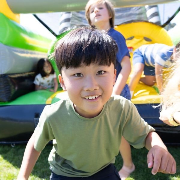A young boy in a green shirt smiles at the camera while playing with other children on an inflatable bounce house outdoors.