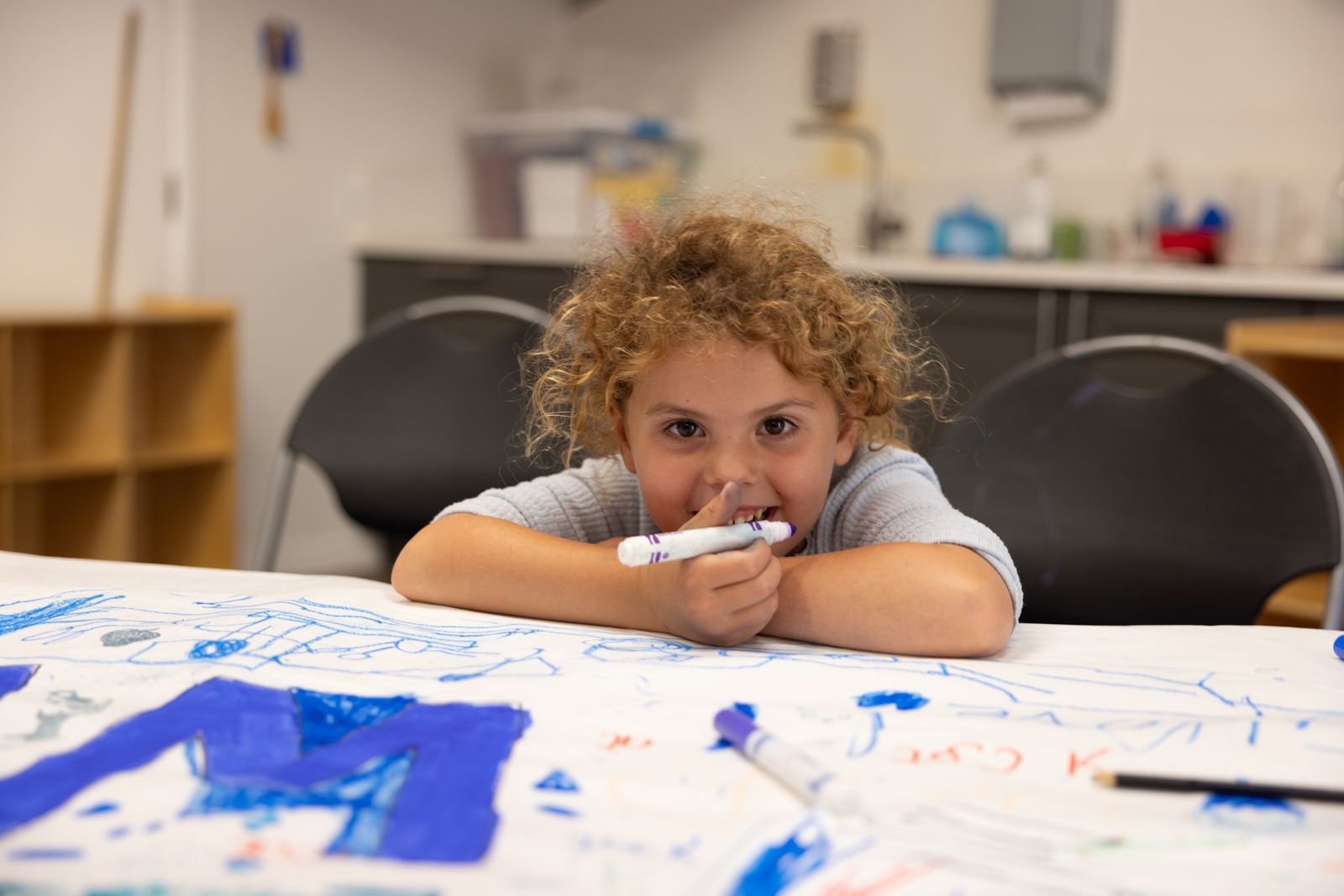 A child with curly hair sits at a table covered in drawings, holding markers and looking at the camera in a classroom setting.