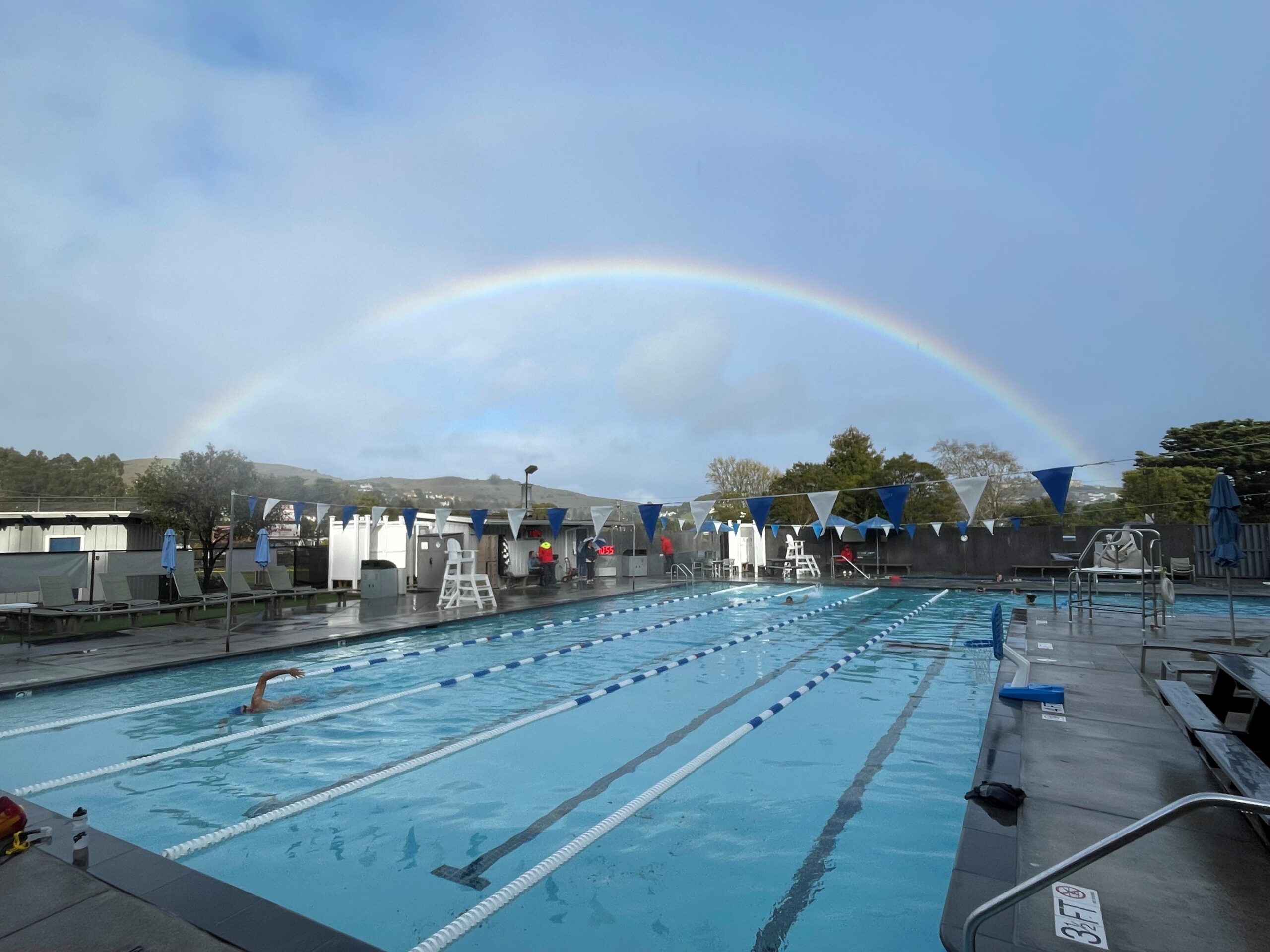 Outdoor swimming pool with lane dividers and lifeguard chairs, under a cloudy sky with a visible rainbow arching overhead.