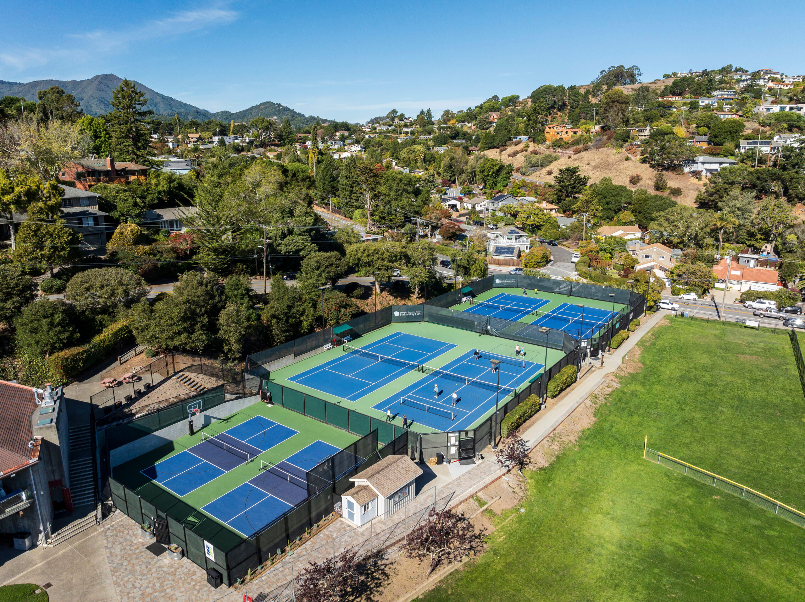 Aerial view of multiple outdoor tennis and pickleball courts next to a grassy field and residential neighborhood on a clear day.