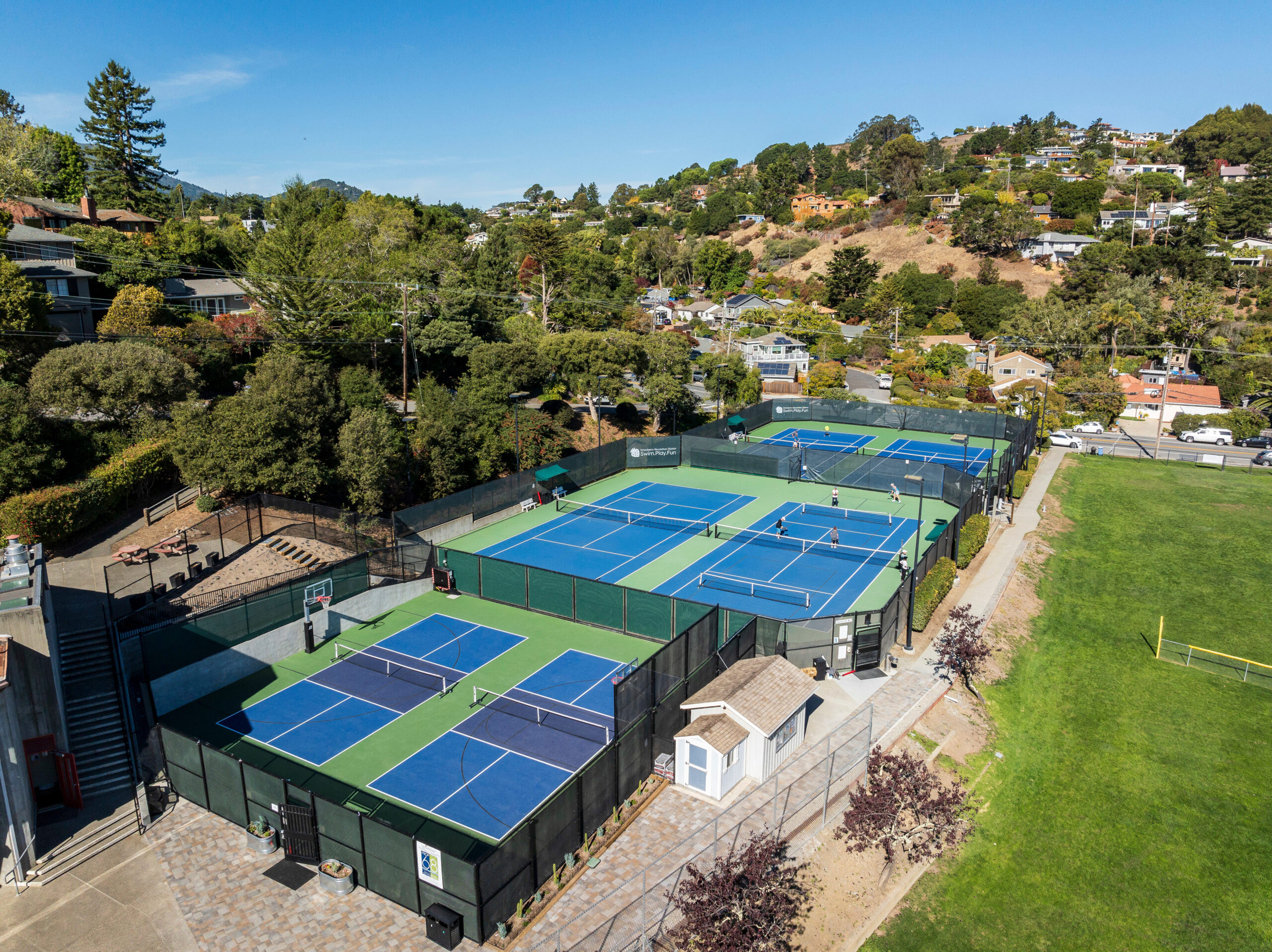 Aerial view of a tennis facility with multiple blue and green courts, surrounded by fencing, near a grassy field and residential neighborhood on a sunny day.