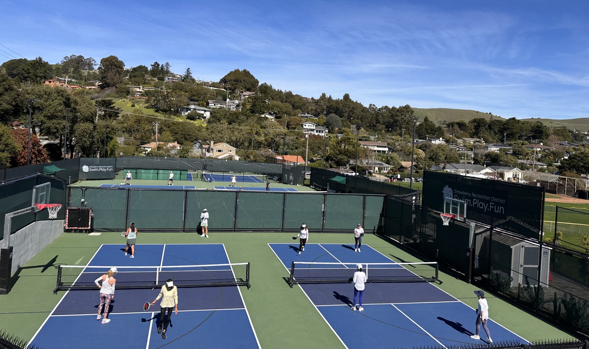 Several people play pickleball on outdoor courts surrounded by green fencing, with houses and trees visible in the background under a clear sky.