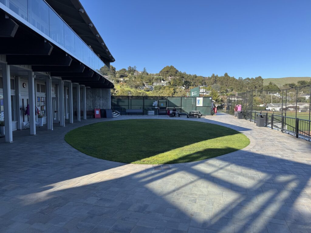 A circular grassy area surrounded by stone pavement next to a building and fenced sports field under a clear blue sky.