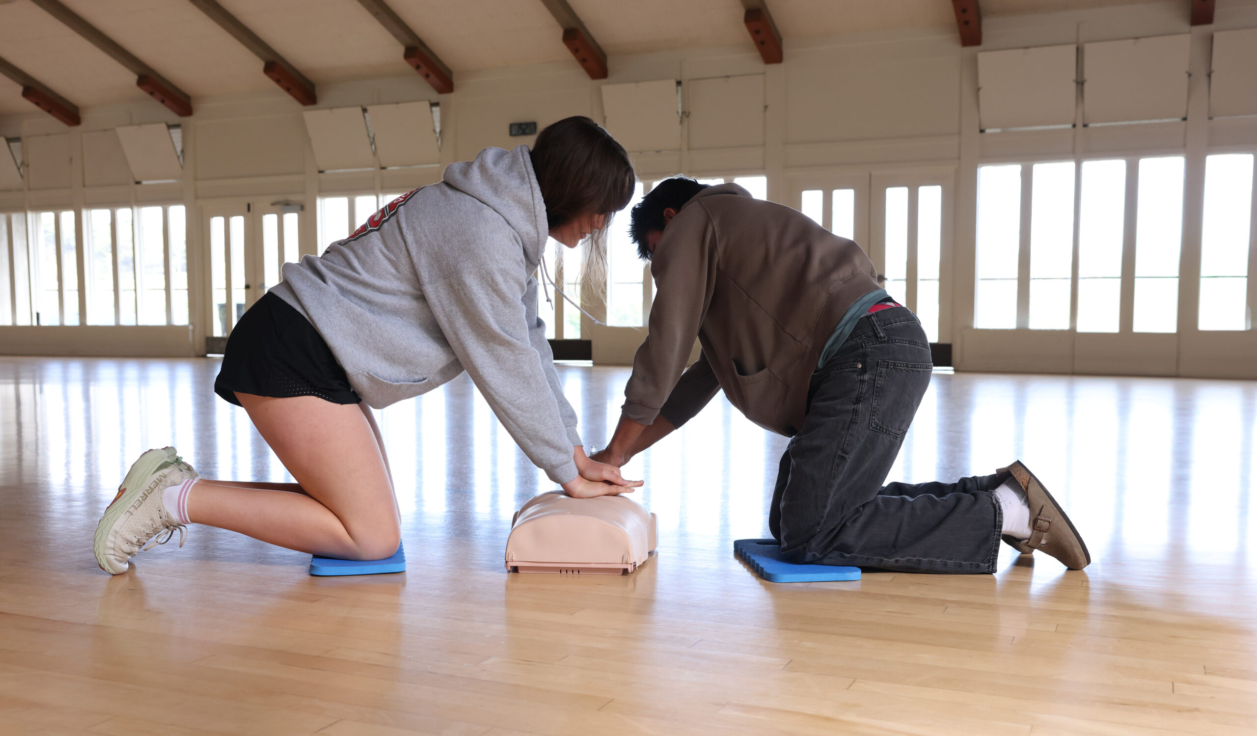 Two people kneeling on mats practice CPR chest compressions on a training mannequin in a bright, spacious room.