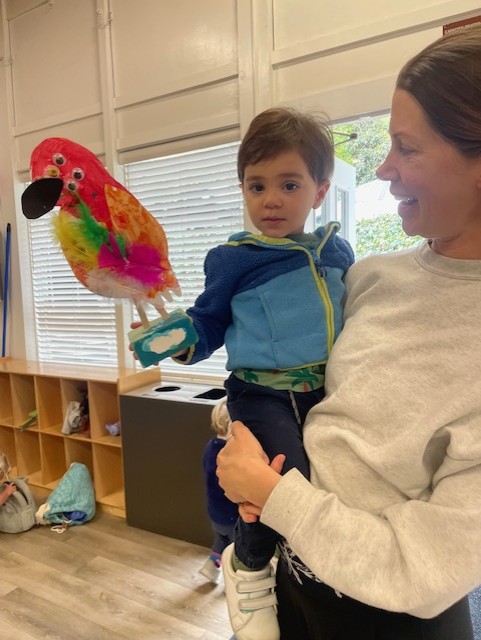 A woman holds a young child indoors; the child is holding a colorful parrot puppet. Shelves and a window are visible in the background.