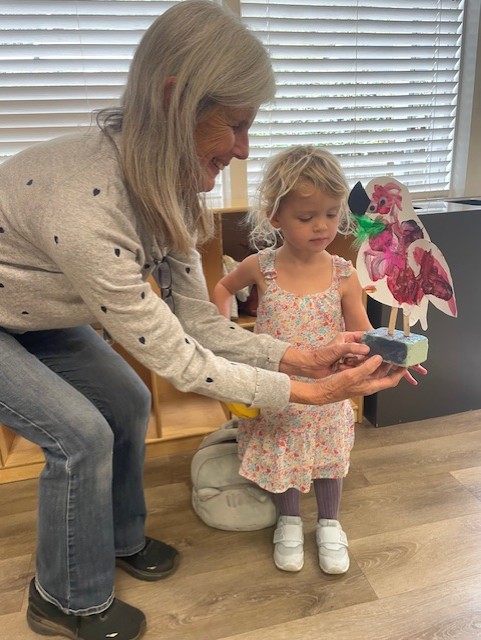 An older woman and a young girl hold a small craft project shaped like a bird in a room with wooden floors and window blinds.