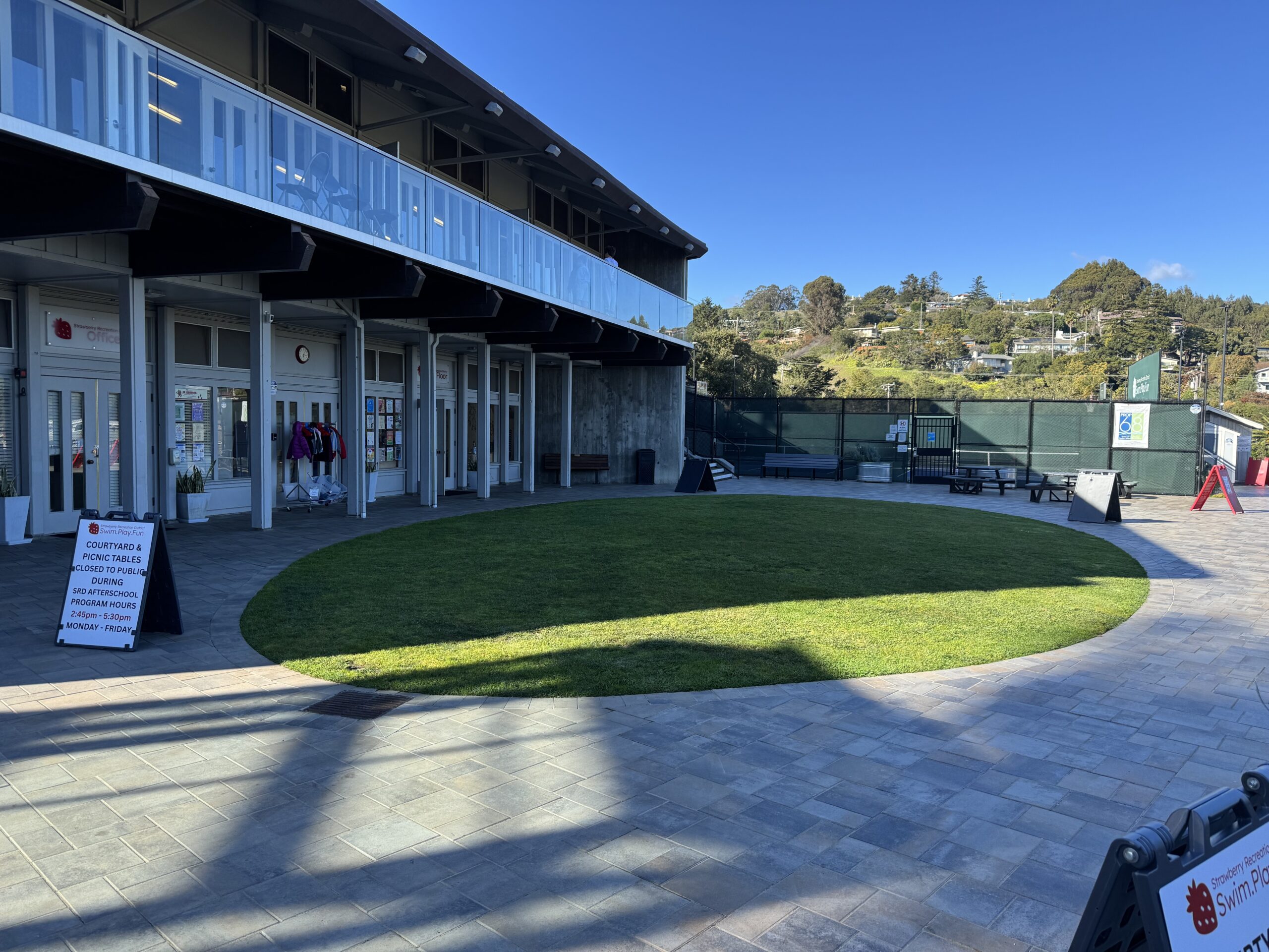 A modern building with glass doors opens onto a circular lawn and paved courtyard, with signs and benches visible under clear, sunny skies.