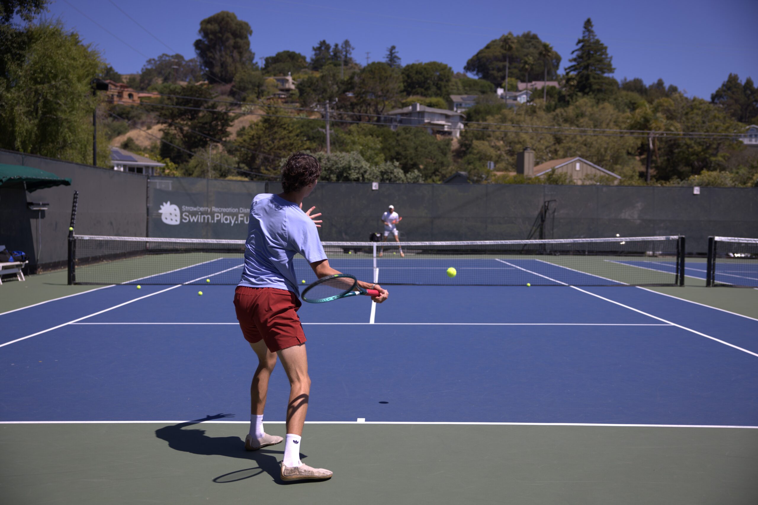 Two people play tennis on an outdoor court; one is preparing to hit the ball while the other stands ready on the opposite side.