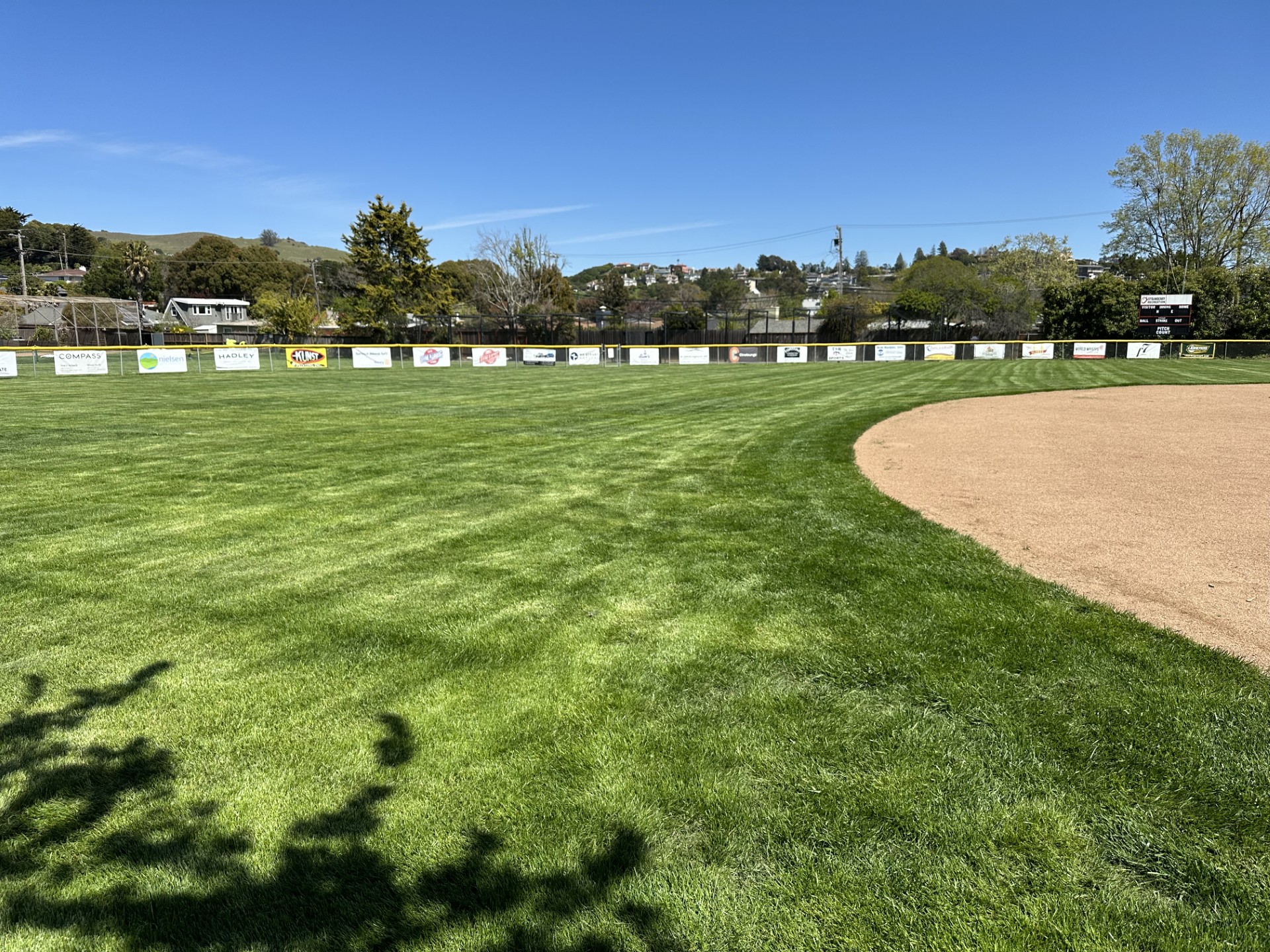 A baseball outfield with a grassy field, dirt warning track, and advertisements on the outfield fence under a clear blue sky.