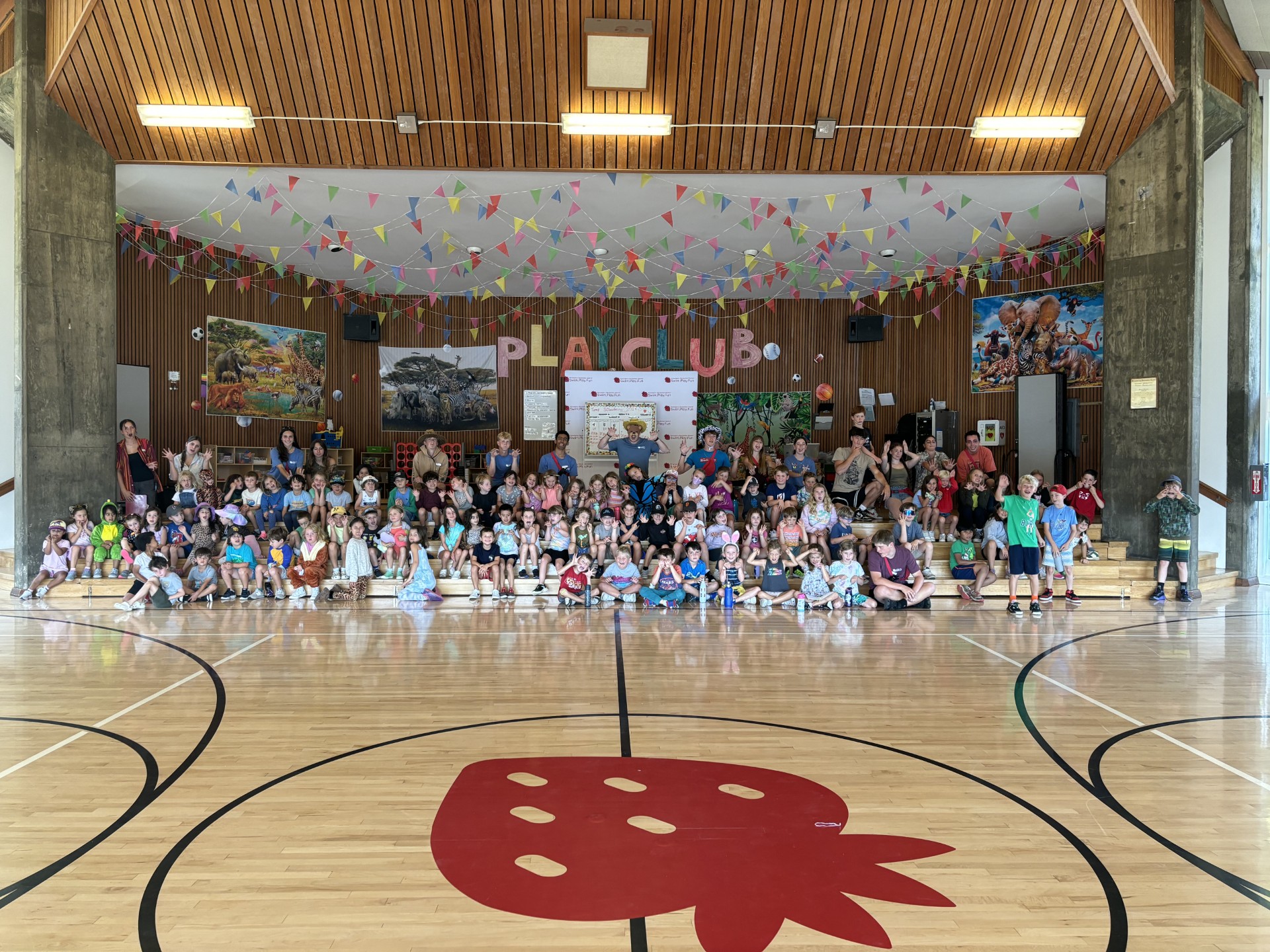 A large group of children and adults sit and stand in a gymnasium decorated with bunting and a “PLAY CLUB” sign, posed for a group photo on a wooden floor with a red strawberry graphic.