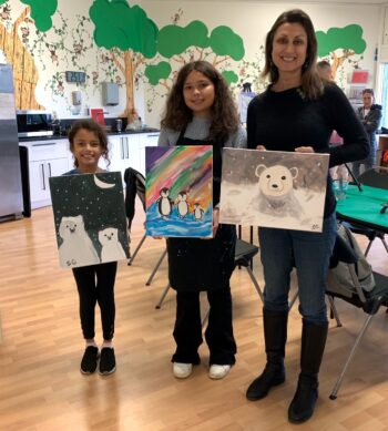 Three people stand indoors holding up paintings of polar bears and penguins; there are tables, chairs, and wall art of trees in the background.