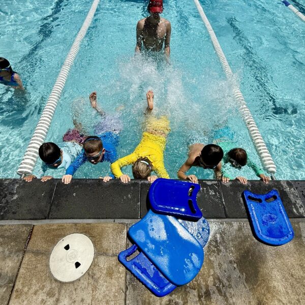Children in swimwear practice kicking at the edge of a pool, supervised by an instructor in a red cap; blue kickboards are on the poolside.
