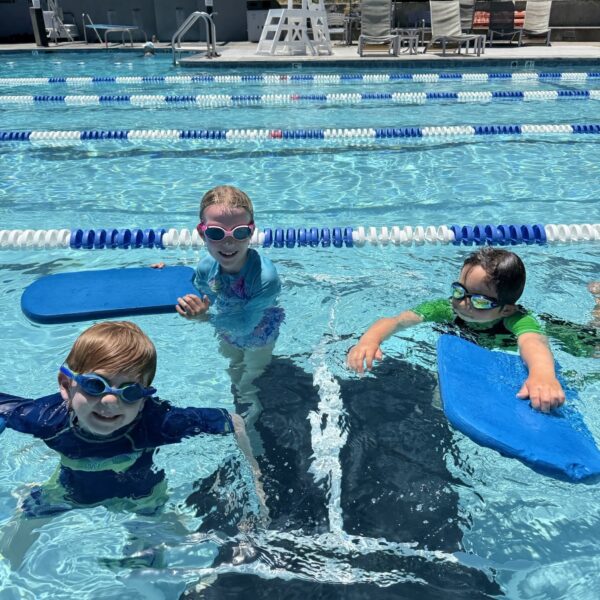 Three children wearing swim goggles and rash guards hold kickboards while standing in a swimming pool, with empty pool chairs and umbrellas in the background.