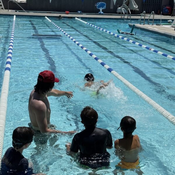 A group of children in swim caps and a swim instructor watch a child practice kicking in a lane of an outdoor swimming pool.