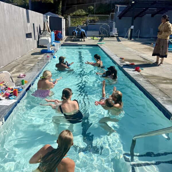 A group of children and adults participate in a swim class in an outdoor pool while an instructor stands at the poolside.