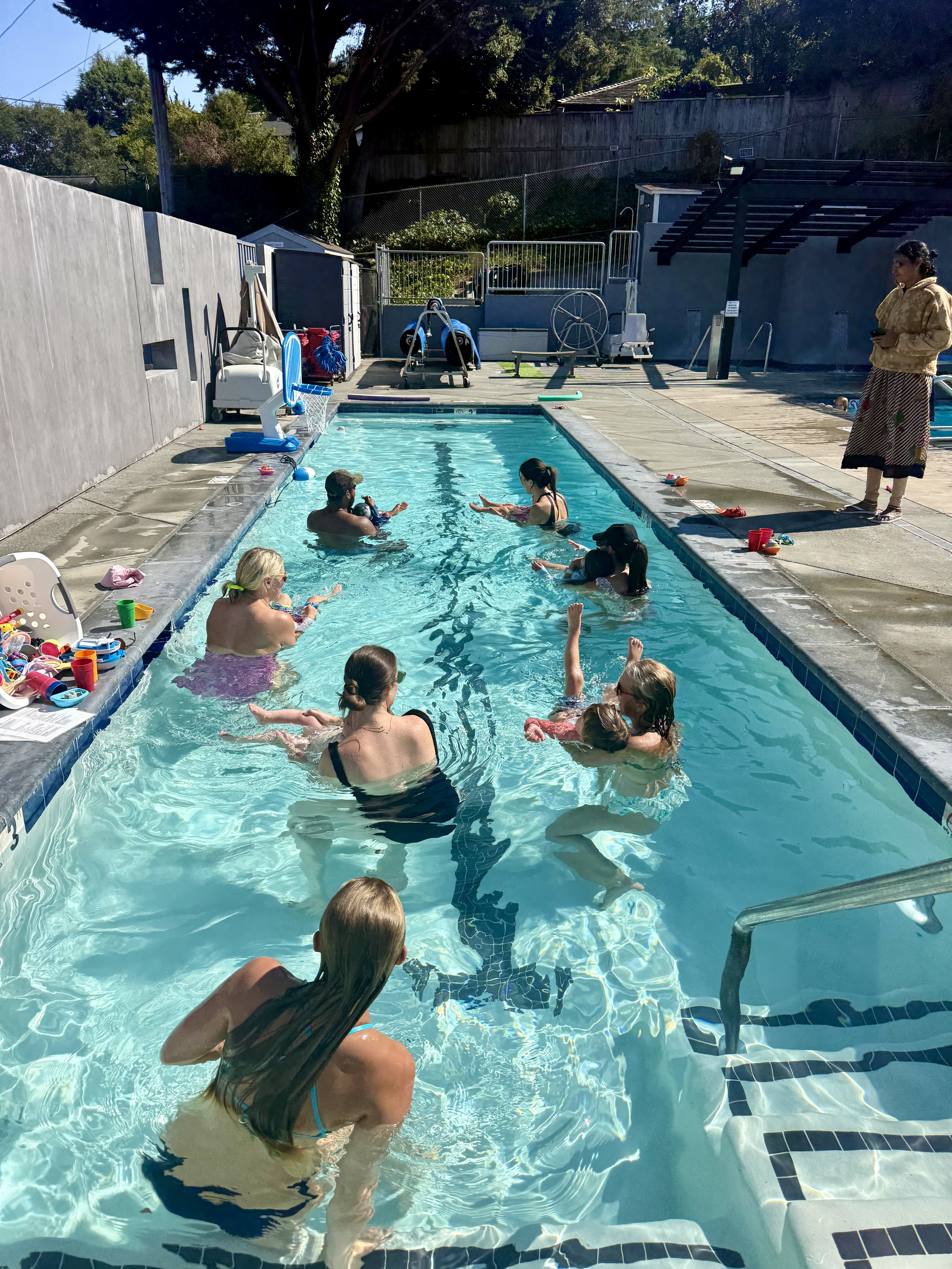 A group of children and adults participate in a swim class in an outdoor pool while an instructor stands at the poolside.