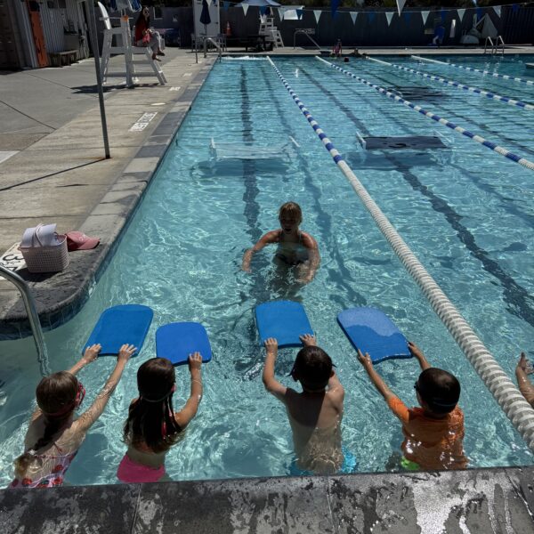 A swim instructor in a pool teaches five young children holding kickboards at the edge of a swim lane on a sunny day.